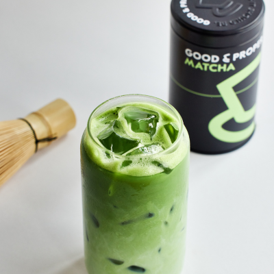 A vibrant green, creamy Iced Matcha Latte sits on a grey background with a bamboo matcha whisk and Good & Proper Matcha Tin-For-Life