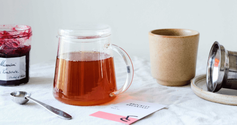 A pot of tea with a metal spoon, fruit preserve and brown cup surrounding it on a table