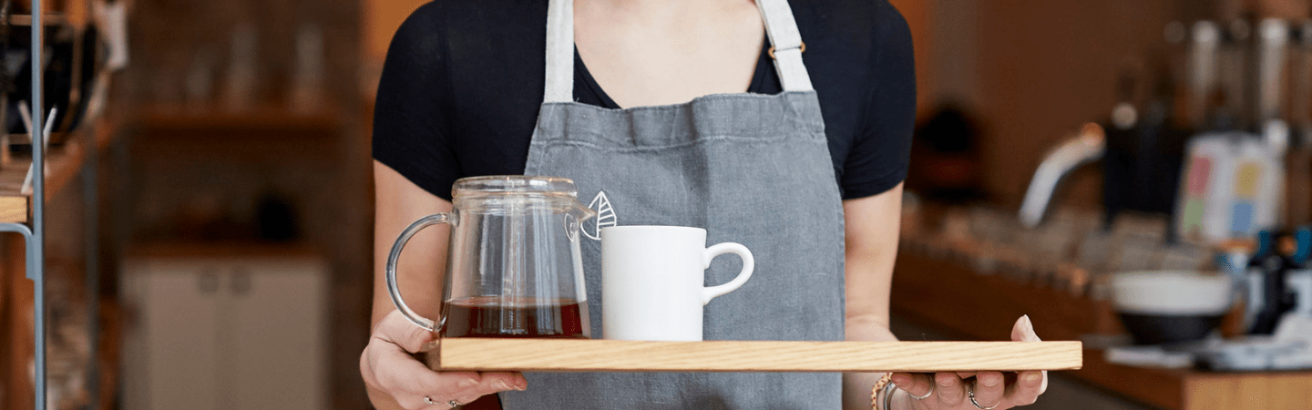 A person in a black top and denim apron carrying a mug of tea on a tray through a cafe