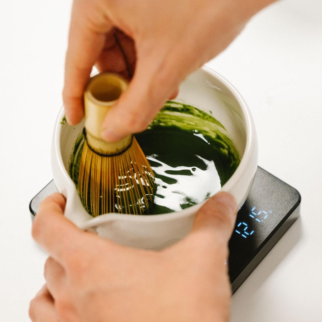 Person using a whisk to mix green tea in a white bowl on a digital scale.