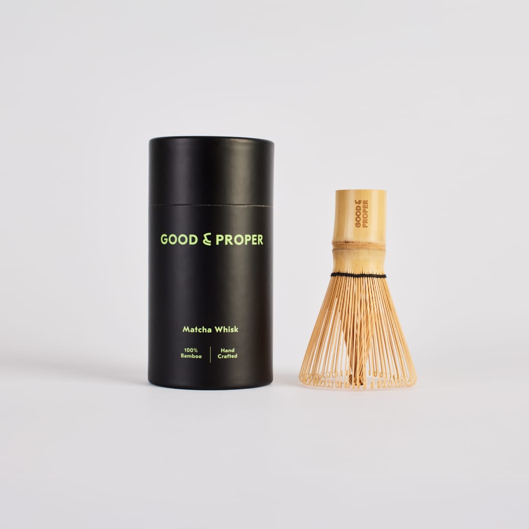 Black cylindrical container with 'Good & Proper' branding next to a wooden matcha whisk on a light gray background