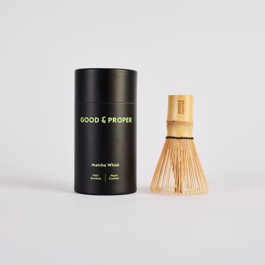 Black cylindrical container with 'Good & Proper' branding next to a wooden matcha whisk on a light gray background