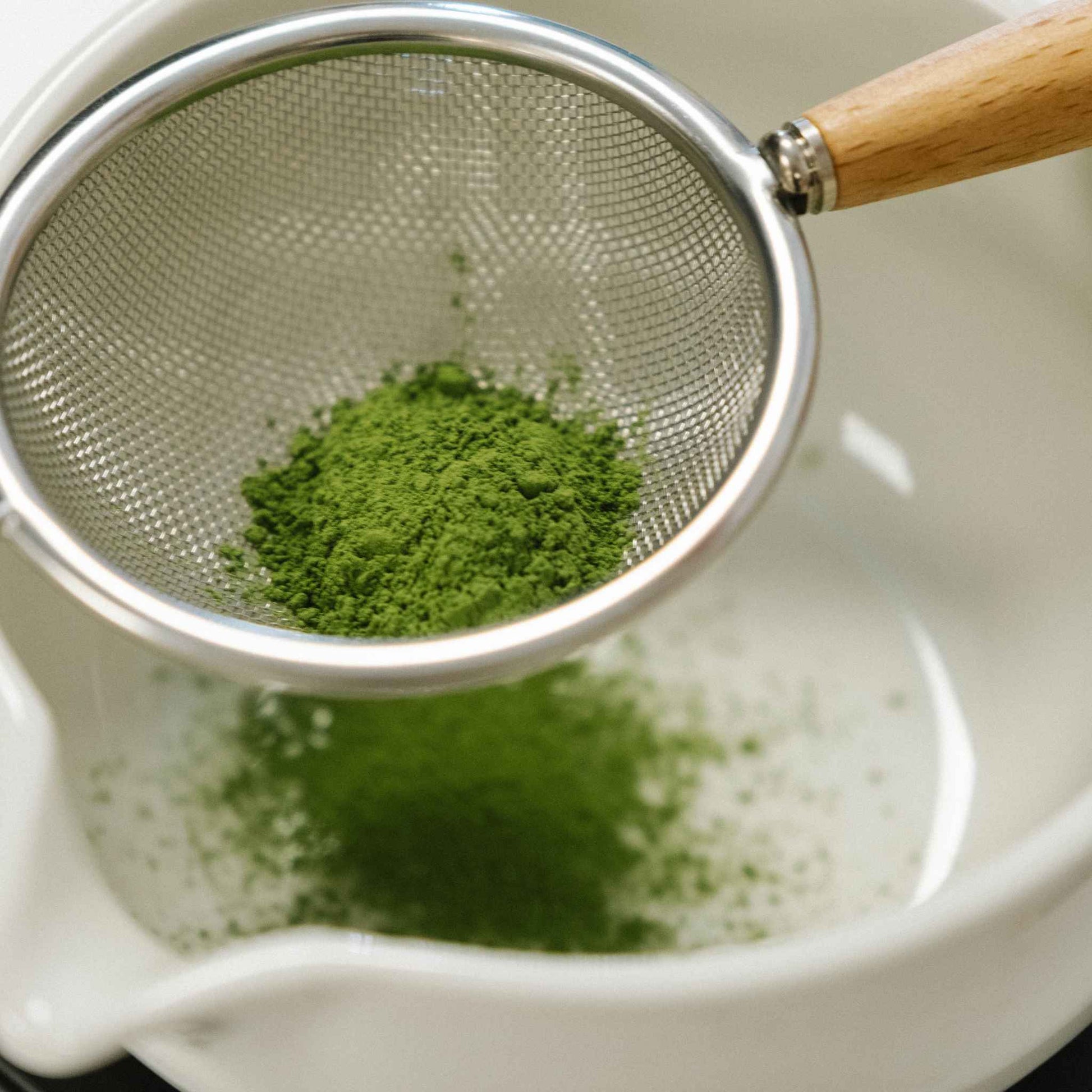 Signature Matcha Green powder in a metal sieve over a white bowl